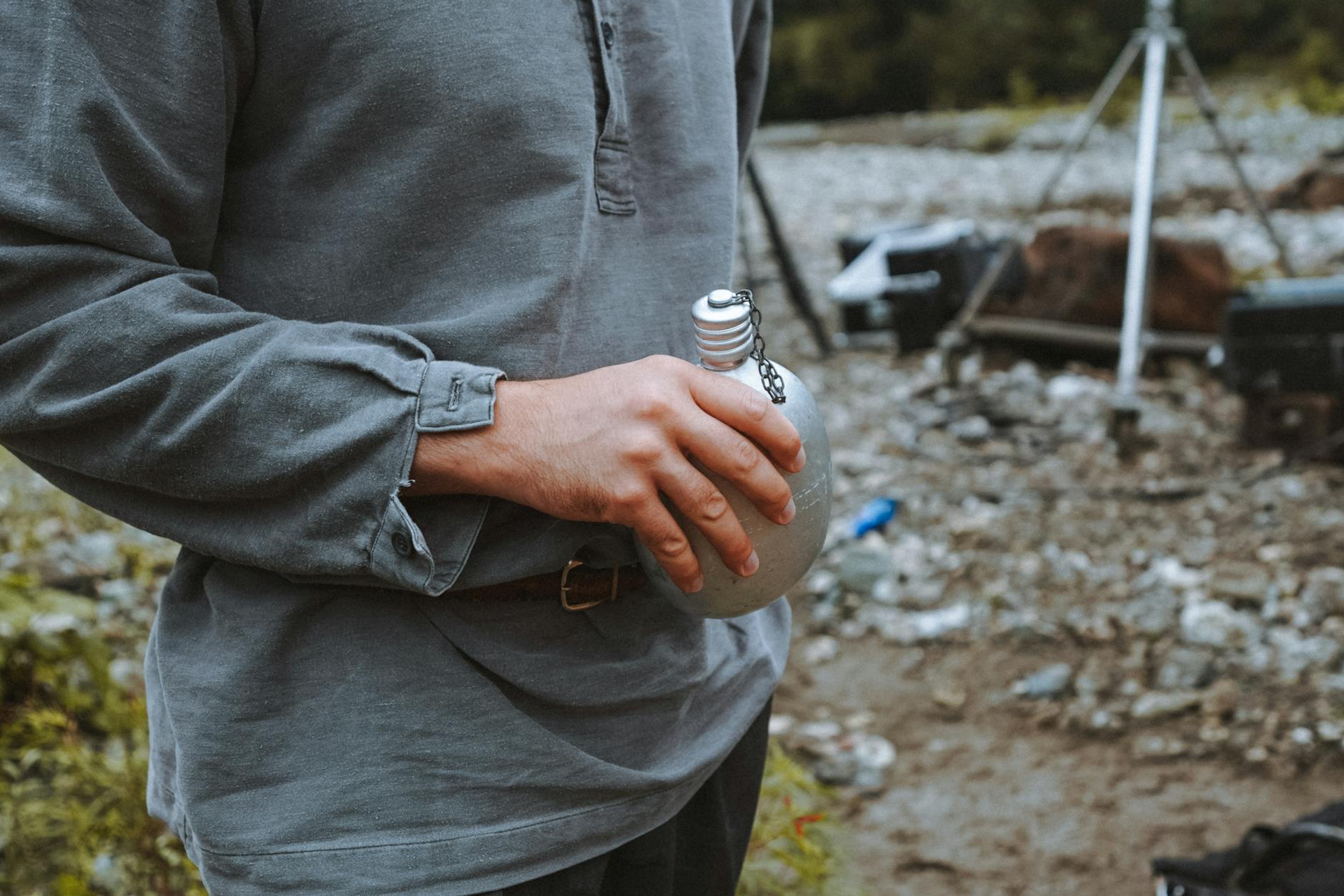 Casual scene of a man holding a tin bottle at a rocky outdoor campsite. Adventure vibe.