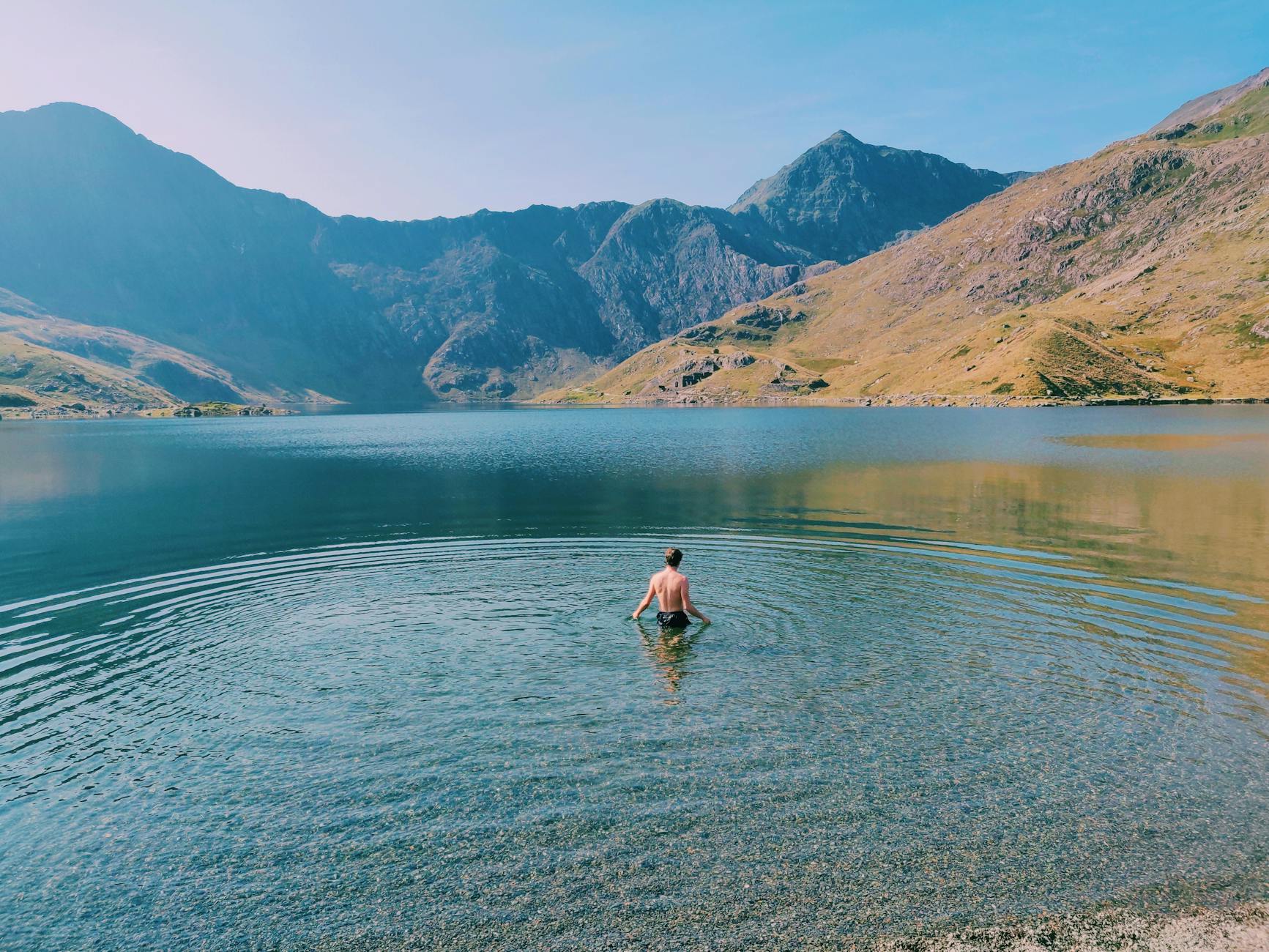 A man swimming in a tranquil lake in Wales, surrounded by scenic mountain views at sunrise.