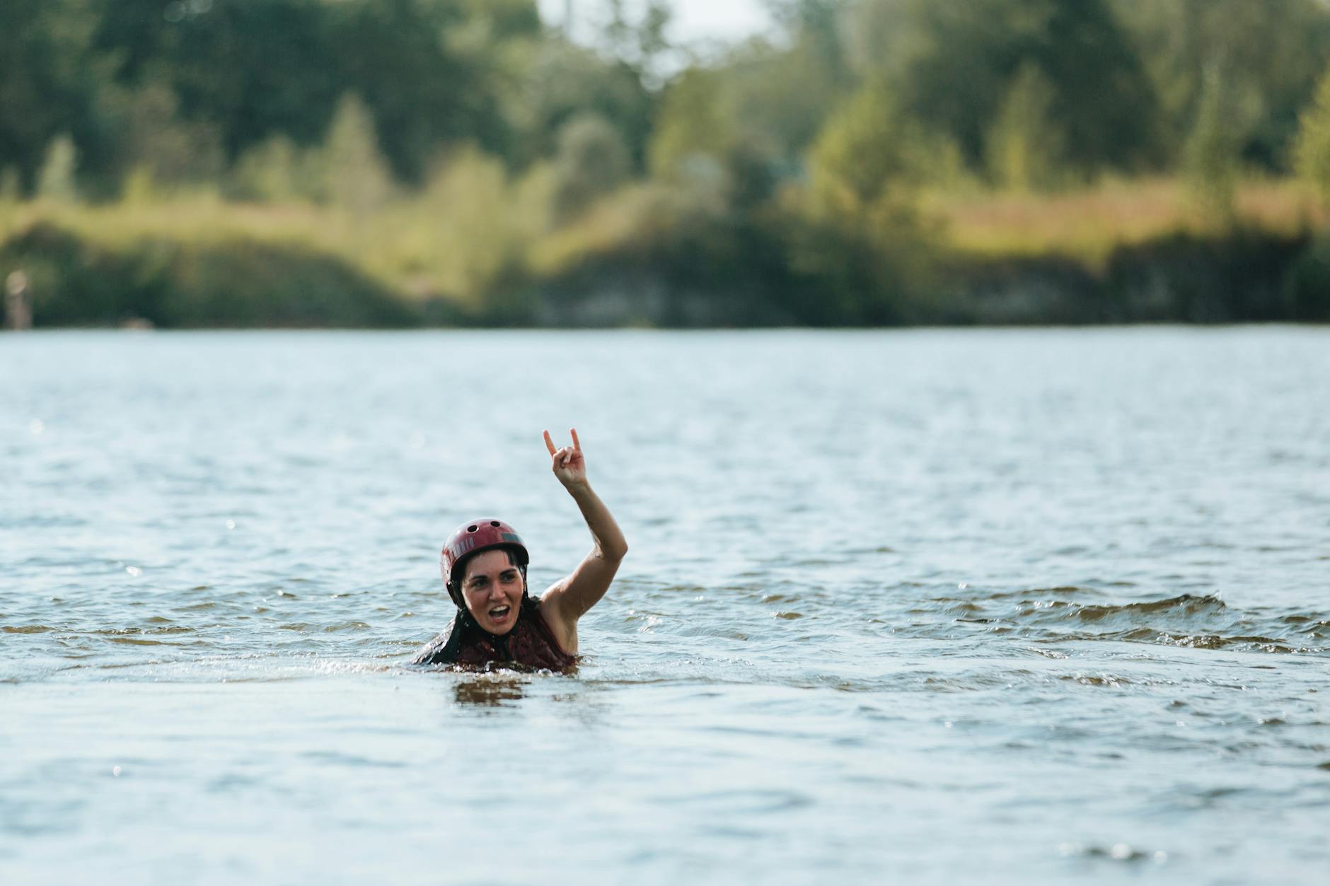 A woman wearing a helmet swims in a river, raising her hand in a gesture with a scenic outdoor background.